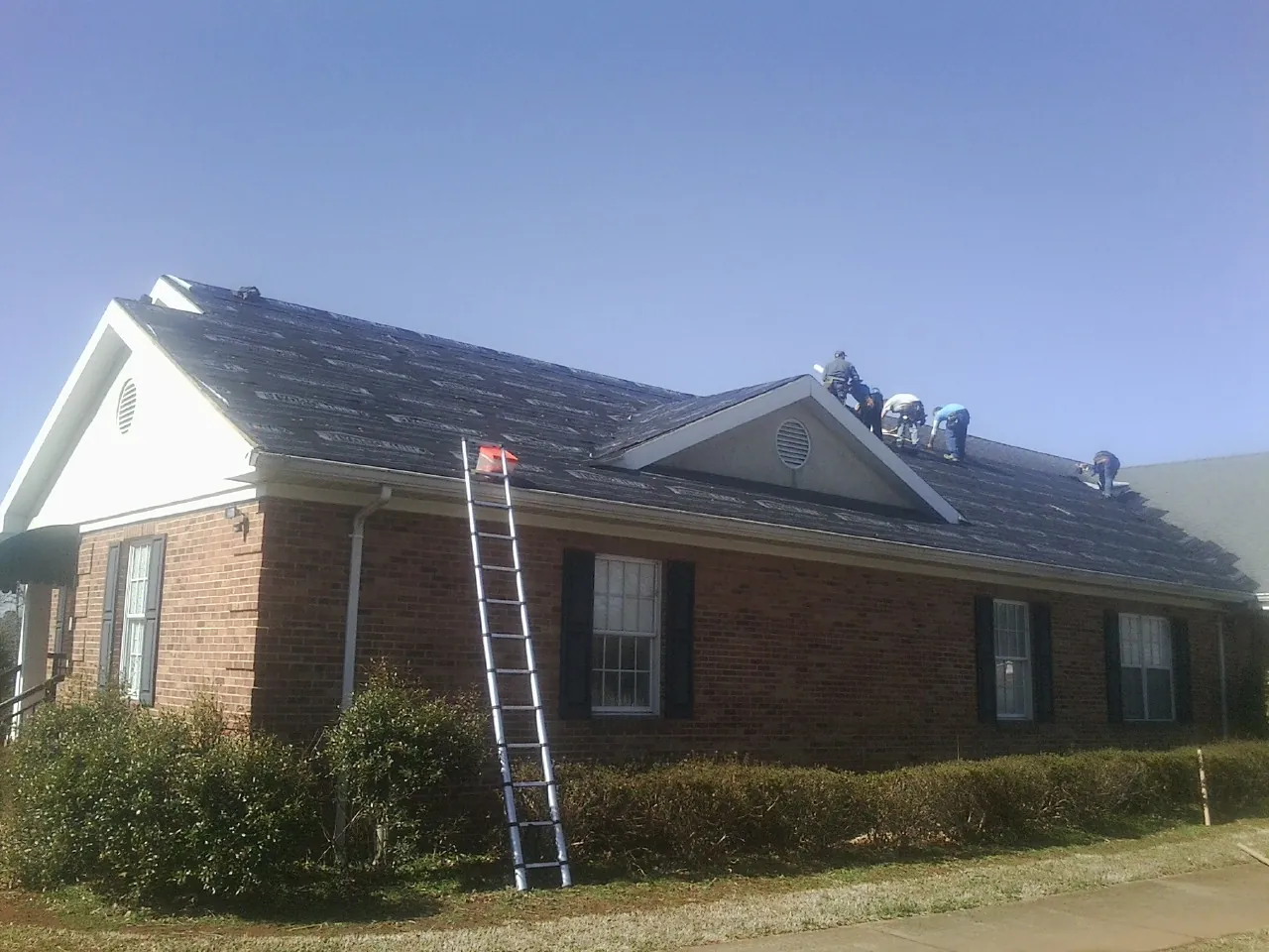 Crew tearing shingles off a brick ranch roof with a ladder set — Metal Roof Repair work by our crew in Wilsons Mills