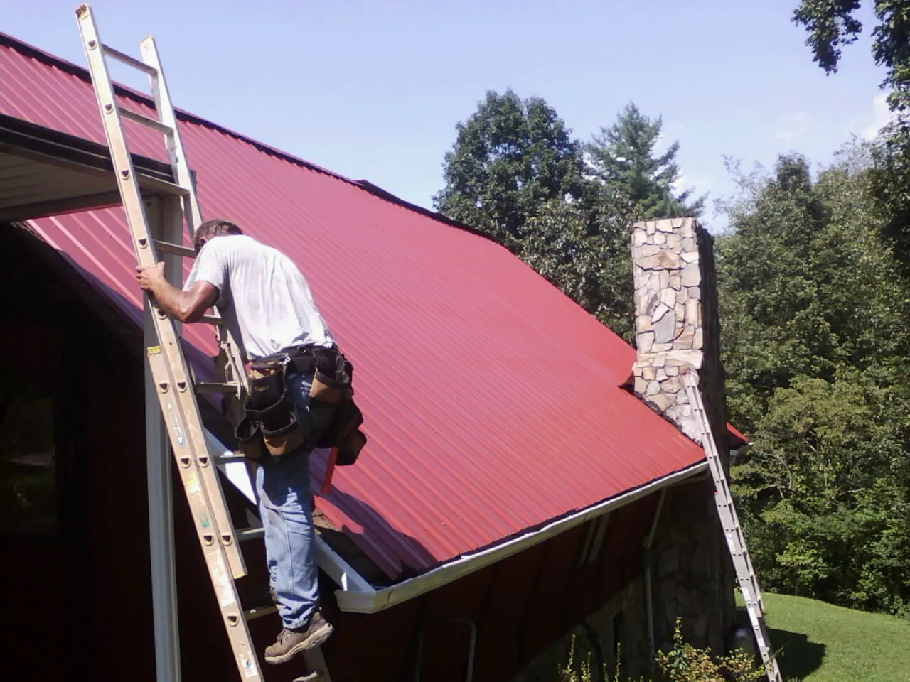 Roofer on a ladder beside a red metal roof and stone chimney — Soffit & Fascia Repair by Roofing Brothers in Wilsons Mills